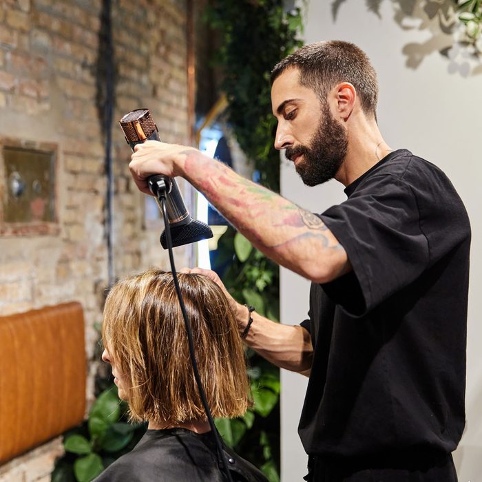 A stylist blow-drying a client&rsquo;s hair in a small salon