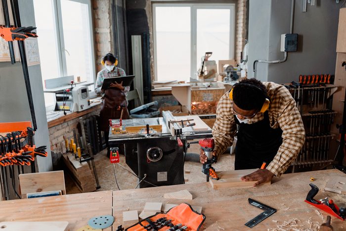 A carpenter marking a board while a coworker works behind them