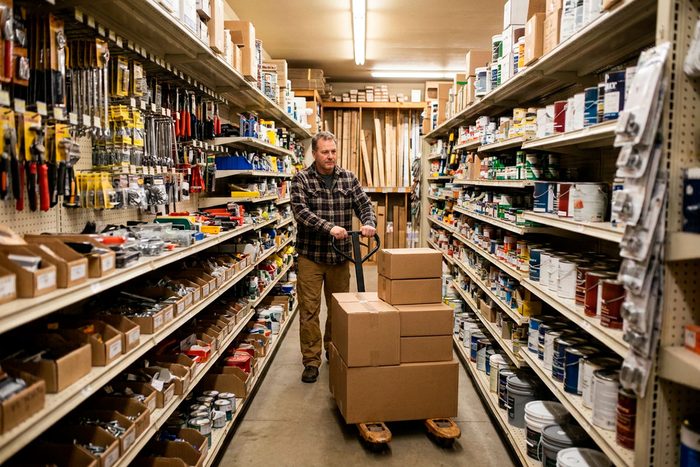 A hardware shop owner wheels a pallet of newly delivered stock down an aisle lined with paint and tools.