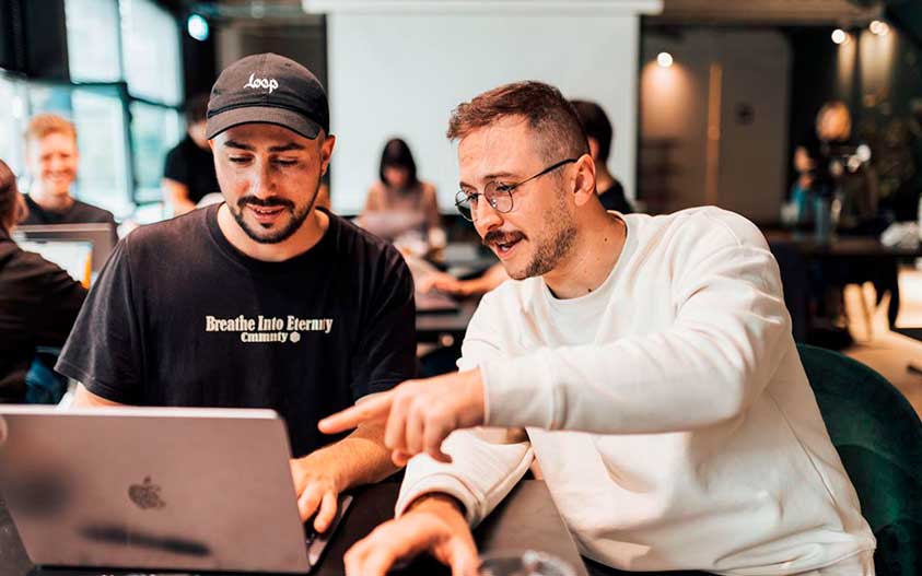 A Cardiff team member at a shared work table, colleagues working at desks behind them.